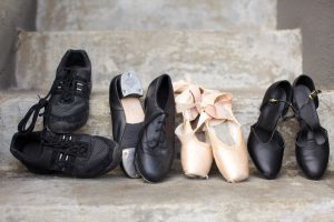 Closeup of a pair of jazz shoes, tap shoes, ballet pointe shoes, and character shoes representing a variety of dance classes in one image. Shoes are resting on concrete steps.